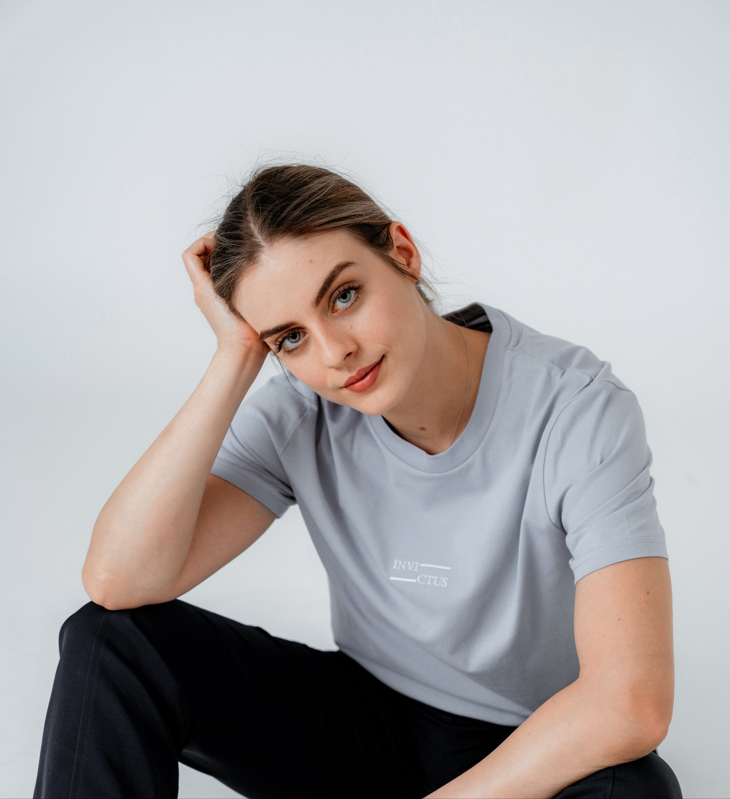 Woman wearing a light gray t-shirt with the invictus sportswear ogo, sitting against a plain background